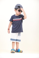 A young child wearing a navy blue t-shirt with a red stripe, light shorts with blue trim, colorful Crocs, and a blue and white striped cap worn backwards is adjusting their sunglasses. The child stands against a plain light background, with one hand touching the sunglasses and the other hanging by their side.