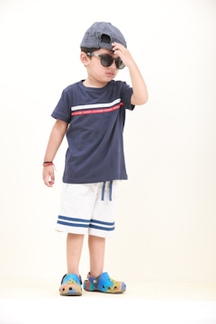 A young child wearing a navy blue t-shirt with a red stripe, light shorts with blue trim, colorful Crocs, and a blue and white striped cap worn backwards is adjusting their sunglasses. The child stands against a plain light background, with one hand touching the sunglasses and the other hanging by their side.