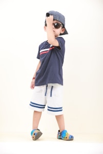 A young child is posing with confidence, wearing a navy blue t-shirt, white shorts with blue stripes, colorful crocs, sunglasses, and a striped cap worn backward. The child is slightly tilting their head down while adjusting the cap with one hand, conveying a playful and cool demeanor. The background is a plain white, emphasizing the subject.