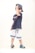 A young child is posing with confidence, wearing a navy blue t-shirt, white shorts with blue stripes, colorful crocs, sunglasses, and a striped cap worn backward. The child is slightly tilting their head down while adjusting the cap with one hand, conveying a playful and cool demeanor. The background is a plain white, emphasizing the subject.