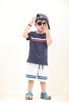 A young child stands confidently wearing a navy blue t-shirt with a red and white stripe, light gray shorts with blue stripes, and colorful Crocs. The child accessorizes with sunglasses and a cap worn backwards, striking a playful pose.