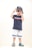 A young child stands confidently wearing a navy blue t-shirt with a red and white stripe, light gray shorts with blue stripes, and colorful Crocs. The child accessorizes with sunglasses and a cap worn backwards, striking a playful pose.