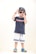 A young child stands confidently wearing a navy blue t-shirt with a red and white stripe, light gray shorts with blue stripes, and colorful Crocs. The child accessorizes with sunglasses and a cap worn backwards, striking a playful pose.