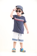 A young child is wearing a navy blue t-shirt with a red and white stripe design, paired with white shorts that have blue stripes at the bottom. The child is accessorized with a striped blue and white cap worn backward, sunglasses, and colorful slip-on shoes. The child is striking a pose with one hand adjusting the cap, exuding a sense of confidence.
