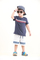 A young child is wearing a navy blue t-shirt with a red and white stripe design, paired with white shorts that have blue stripes at the bottom. The child is accessorized with a striped blue and white cap worn backward, sunglasses, and colorful slip-on shoes. The child is striking a pose with one hand adjusting the cap, exuding a sense of confidence.