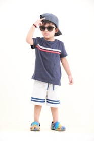 A young child is wearing a navy blue t-shirt with a red and white stripe design, paired with white shorts that have blue stripes at the bottom. The child is accessorized with a striped blue and white cap worn backward, sunglasses, and colorful slip-on shoes. The child is striking a pose with one hand adjusting the cap, exuding a sense of confidence.