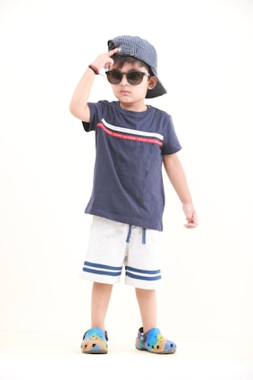 A young child is wearing a navy blue t-shirt with a red and white stripe design, paired with white shorts that have blue stripes at the bottom. The child is accessorized with a striped blue and white cap worn backward, sunglasses, and colorful slip-on shoes. The child is striking a pose with one hand adjusting the cap, exuding a sense of confidence.