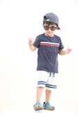A child wearing a navy blue t-shirt with red and white accents, white shorts with blue stripes, and colorful clog shoes. The child is also wearing sunglasses and a striped cap turned backwards, standing against a plain background.