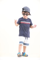 A child wearing a navy blue t-shirt with red and white accents, white shorts with blue stripes, and colorful clog shoes. The child is also wearing sunglasses and a striped cap turned backwards, standing against a plain background.