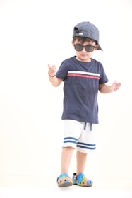 A child wearing a navy blue t-shirt with red and white accents, white shorts with blue stripes, and colorful clog shoes. The child is also wearing sunglasses and a striped cap turned backwards, standing against a plain background.