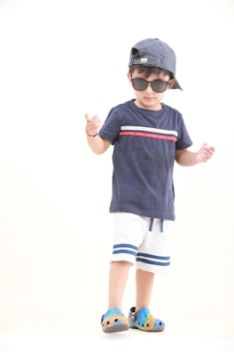 A child wearing a navy blue t-shirt with red and white accents, white shorts with blue stripes, and colorful clog shoes. The child is also wearing sunglasses and a striped cap turned backwards, standing against a plain background.