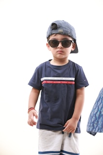 A young child is wearing large black sunglasses and a striped blue cap worn backwards. The child is dressed in a navy blue T-shirt with white and red stripes on the chest, and light-colored shorts with a blue band near the bottom. The child has a calm expression and stands against a plain light background.