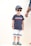 A young child is wearing a blue t-shirt with a red and white stripe pattern across the chest, paired with white shorts featuring blue stripes at the bottom. The child has on a blue baseball cap and dark sunglasses, and is standing with a neutral expression. Colorful clogs featuring blue and yellow hues are on the feet. An adult's hand, wearing bracelets, is partially visible to the right.