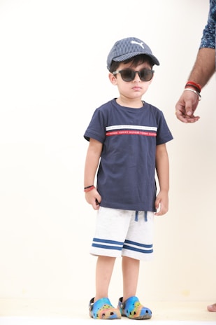 A young child is wearing a blue t-shirt with a red and white stripe pattern across the chest, paired with white shorts featuring blue stripes at the bottom. The child has on a blue baseball cap and dark sunglasses, and is standing with a neutral expression. Colorful clogs featuring blue and yellow hues are on the feet. An adult's hand, wearing bracelets, is partially visible to the right.