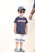 A young child is wearing a blue t-shirt with a red and white stripe pattern across the chest, paired with white shorts featuring blue stripes at the bottom. The child has on a blue baseball cap and dark sunglasses, and is standing with a neutral expression. Colorful clogs featuring blue and yellow hues are on the feet. An adult's hand, wearing bracelets, is partially visible to the right.