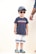 A young child is wearing a blue t-shirt with a red and white stripe pattern across the chest, paired with white shorts featuring blue stripes at the bottom. The child has on a blue baseball cap and dark sunglasses, and is standing with a neutral expression. Colorful clogs featuring blue and yellow hues are on the feet. An adult's hand, wearing bracelets, is partially visible to the right.