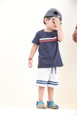 A young child wearing a blue t-shirt and light-colored shorts, with blue and green shoes. The child is wearing a striped cap and sunglasses, covering part of their face with their hand. An adult hand is visible near the child.