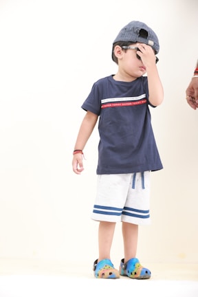 A young child wearing a blue t-shirt and light-colored shorts, with blue and green shoes. The child is wearing a striped cap and sunglasses, covering part of their face with their hand. An adult hand is visible near the child.