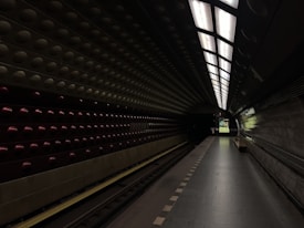 A dimly lit underground subway station with a modern and sleek design. The walls are adorned with circular patterns and dark tiles. The platform is empty, creating a sense of solitude. Overhead lights cast a minimal amount of illumination, contributing to the moody atmosphere.