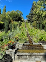 A garden landscape features a cascade of water descending a series of stone steps, surrounded by vibrant greenery and colorful flowers. Tall, lush trees frame the scene against a bright blue sky, creating a serene and picturesque environment.