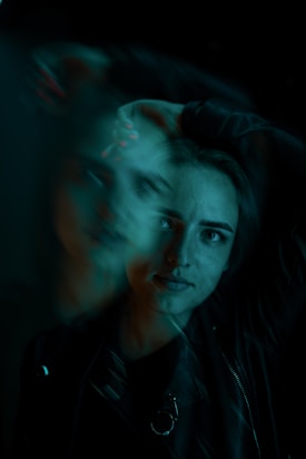 A portrait of a person with a ghostly, motion-blur effect. The subject has one arm raised behind their head and is wearing a dark outfit. The lighting is predominantly teal, creating an enigmatic and artistic atmosphere.