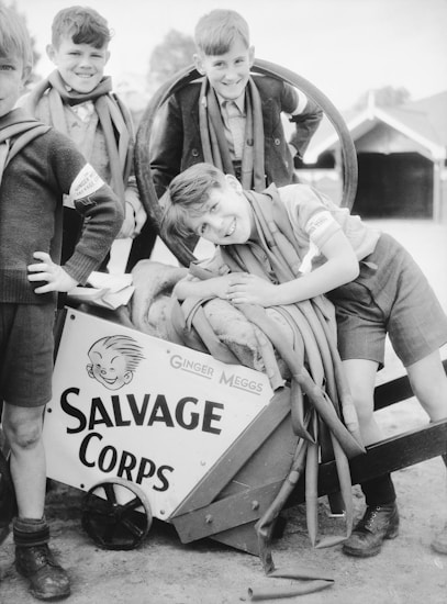 Four boys are playfully gathered around a cart labeled 'SALVAGE CORPS' with a cartoon character named Ginger Meggs. They are smiling and dressed in old-fashioned clothing, with hoses draped over their shoulders.