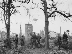 A group of soldiers in helmets stand in a desolate landscape with barren trees and the ruins of buildings. In the background, a large balloon hovers in the sky, adding to the scene of a war-torn environment.