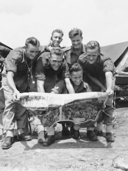 A group of six young men in military uniforms crouches together outdoors. They are closely examining a large newspaper or map, smiling and appearing engaged. Tents are visible in the background, suggesting a camp setting.
