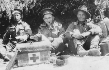 Three soldiers in uniforms are sitting on the ground in an outdoor setting, appearing relaxed. One soldier is smoking a pipe, while another has a cigarette. A medical kit with a red cross is prominently placed on the ground in front of them. They are seated near some foliage, suggesting a campsite or resting spot.