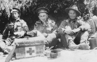 Three soldiers in uniforms are sitting on the ground in an outdoor setting, appearing relaxed. One soldier is smoking a pipe, while another has a cigarette. A medical kit with a red cross is prominently placed on the ground in front of them. They are seated near some foliage, suggesting a campsite or resting spot.