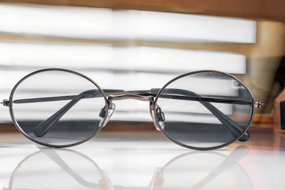 A pair of round, metal-rimmed eyeglasses rests on a reflective surface with a soft focus background consisting of horizontal light bands.