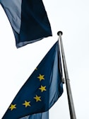 Two European Union flags on poles wave against a cloudy sky. The flags are deep blue with yellow stars arranged in a circle, symbolizing unity.