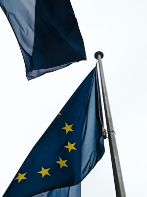 Two European Union flags on poles wave against a cloudy sky. The flags are deep blue with yellow stars arranged in a circle, symbolizing unity.