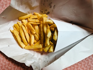 A cone of golden, crispy French fries is placed on white paper, likely as a makeshift plate. The fries are spread out, showcasing a delicious and appetizing look with a slightly oily texture.