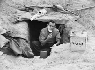 A man is sitting at the entrance of a dugout in a sandy environment, holding a piece of bread. Nearby, there is a container labeled 'WATER'. A tarp covers part of the entrance, and some personal belongings, like a coat and a hat, are visible.
