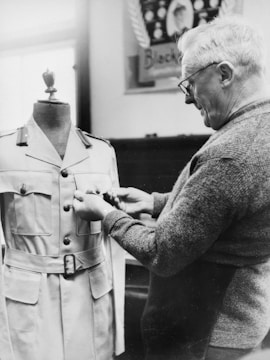 A man is inspecting or adjusting a military uniform on a mannequin. The uniform is neatly buttoned and features a belt. The man, wearing glasses and a sweater, appears to be focused on his task. A blurred poster is visible in the background.
