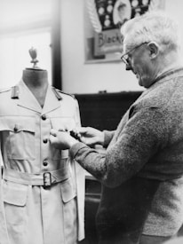 A man is inspecting or adjusting a military uniform on a mannequin. The uniform is neatly buttoned and features a belt. The man, wearing glasses and a sweater, appears to be focused on his task. A blurred poster is visible in the background.