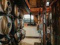 A brewery setup featuring multiple large stainless steel fermentation tanks connected with pipes and tubing. The setting has an industrial feel with brick walls and a wooden ceiling. Natural light filters in through a large arched window in the background, creating a warm and somewhat rustic atmosphere.