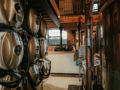 A brewery setup featuring multiple large stainless steel fermentation tanks connected with pipes and tubing. The setting has an industrial feel with brick walls and a wooden ceiling. Natural light filters in through a large arched window in the background, creating a warm and somewhat rustic atmosphere.
