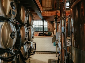 A brewery setup featuring multiple large stainless steel fermentation tanks connected with pipes and tubing. The setting has an industrial feel with brick walls and a wooden ceiling. Natural light filters in through a large arched window in the background, creating a warm and somewhat rustic atmosphere.