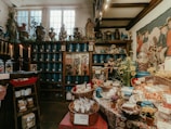 Inside of an old-fashioned shop filled with various goods. In the foreground, baskets of packaged items and candies are displayed on a table with a colorful cloth. Behind the table, wooden shelves line the walls, stocked with blue containers and jars. There are also plants, flowers, and various decorative items on the shelves, creating a vintage and cozy ambiance. Candles are lit on one side, adding to the warm atmosphere.
