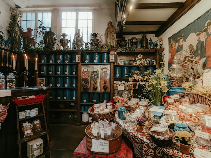 Inside of an old-fashioned shop filled with various goods. In the foreground, baskets of packaged items and candies are displayed on a table with a colorful cloth. Behind the table, wooden shelves line the walls, stocked with blue containers and jars. There are also plants, flowers, and various decorative items on the shelves, creating a vintage and cozy ambiance. Candles are lit on one side, adding to the warm atmosphere.