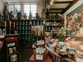 Inside of an old-fashioned shop filled with various goods. In the foreground, baskets of packaged items and candies are displayed on a table with a colorful cloth. Behind the table, wooden shelves line the walls, stocked with blue containers and jars. There are also plants, flowers, and various decorative items on the shelves, creating a vintage and cozy ambiance. Candles are lit on one side, adding to the warm atmosphere.