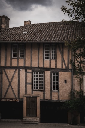 A rustic building with timber-framed construction is depicted under an overcast sky. It features multiple windows and a central wooden door. On the facade, the words 'JOUETS en BOIS' are visible, suggesting a shop or display of wooden toys. The structure has a tiled roof and visible tree branches in the foreground.