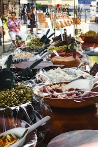 A bustling outdoor market stall displays a wide variety of vibrant antipasti and food items, including olives, cheeses, and spreads. The setup includes wooden bowls and serving utensils. People in the background are casually browsing and walking, creating a lively and inviting atmosphere.
