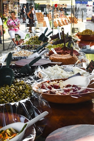 A bustling outdoor market stall displays a wide variety of vibrant antipasti and food items, including olives, cheeses, and spreads. The setup includes wooden bowls and serving utensils. People in the background are casually browsing and walking, creating a lively and inviting atmosphere.
