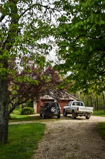 A gravel driveway surrounded by lush greenery leads to a small garage. Two vehicles, a black SUV and a white pickup truck, are parked in front of the garage. Overhanging trees with green leaves create a natural canopy over the scene.