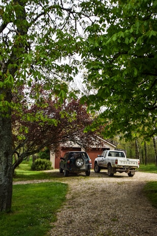 A gravel driveway surrounded by lush greenery leads to a small garage. Two vehicles, a black SUV and a white pickup truck, are parked in front of the garage. Overhanging trees with green leaves create a natural canopy over the scene.