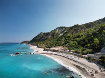 A picturesque coastline with turquoise waters gently lapping against a sandy beach. The beach is lined with lounge chairs and umbrellas, indicating a popular spot for relaxation. Lush green hills rise behind the beach, adding a sense of seclusion and natural beauty to the scene. The sky above is clear and bright, enhancing the vivid colors of the landscape.