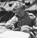A person with light hair concentrates on operating a vintage sewing machine outdoors. The scene includes a fabric piece being worked on, with the sewing machine prominently featuring the brand 'Singer'. The background appears to consist of tents or temporary shelters.